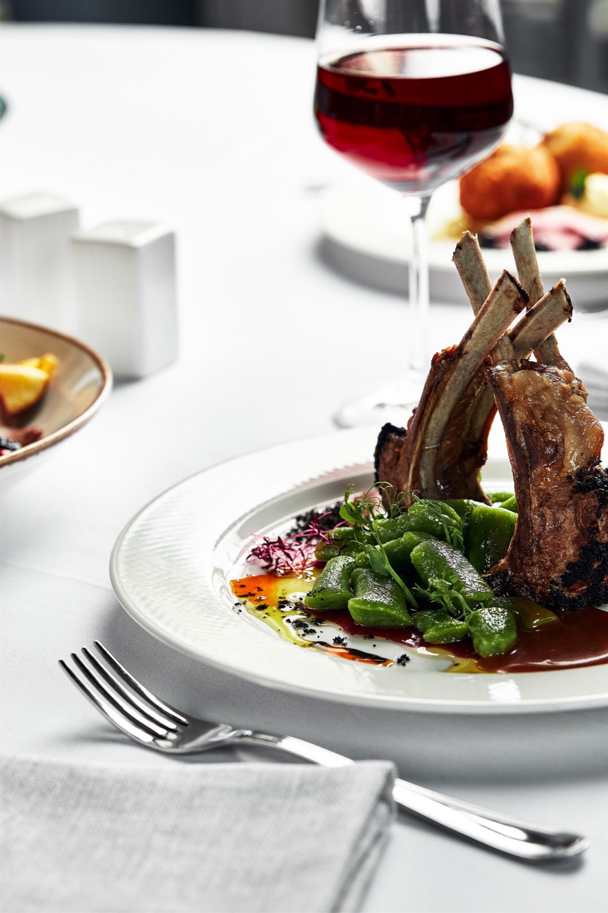 A plated rack of lamb with a glass of red wine on a linen-laid table.