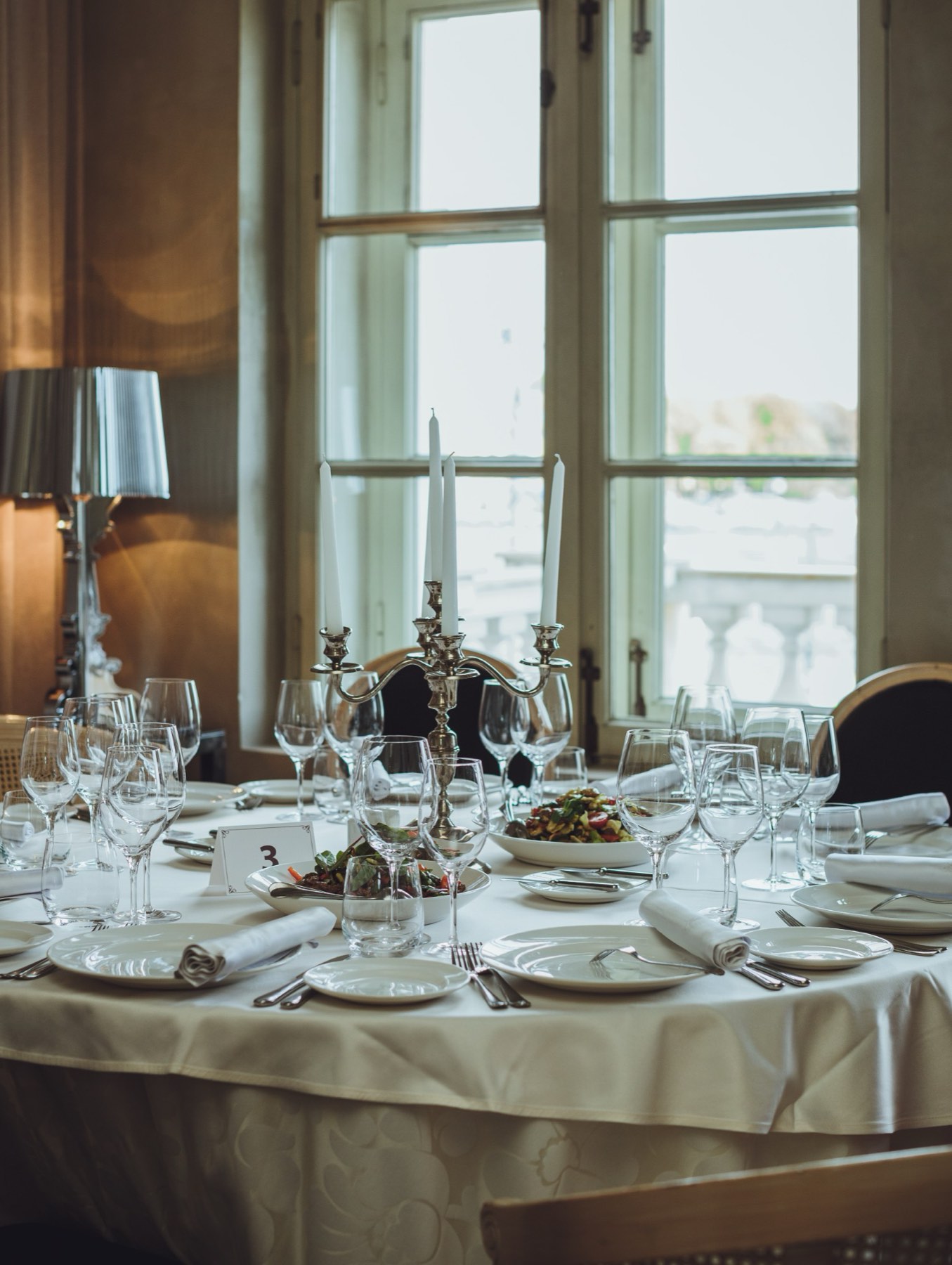 A restaurant table laid by a tall window, with candelabra and glassware.